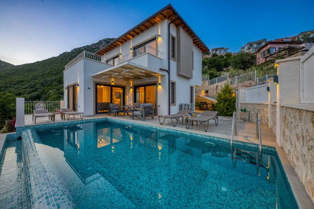 Akşam karanlığında havuzlu iki katlı villa ve dağ manzarası.