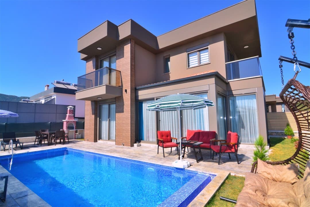 Bahçesinde özel havuzu, şemsiyesi, kırmızı oturma grubu ve salıncağı bulunan iki katlı modern villa.