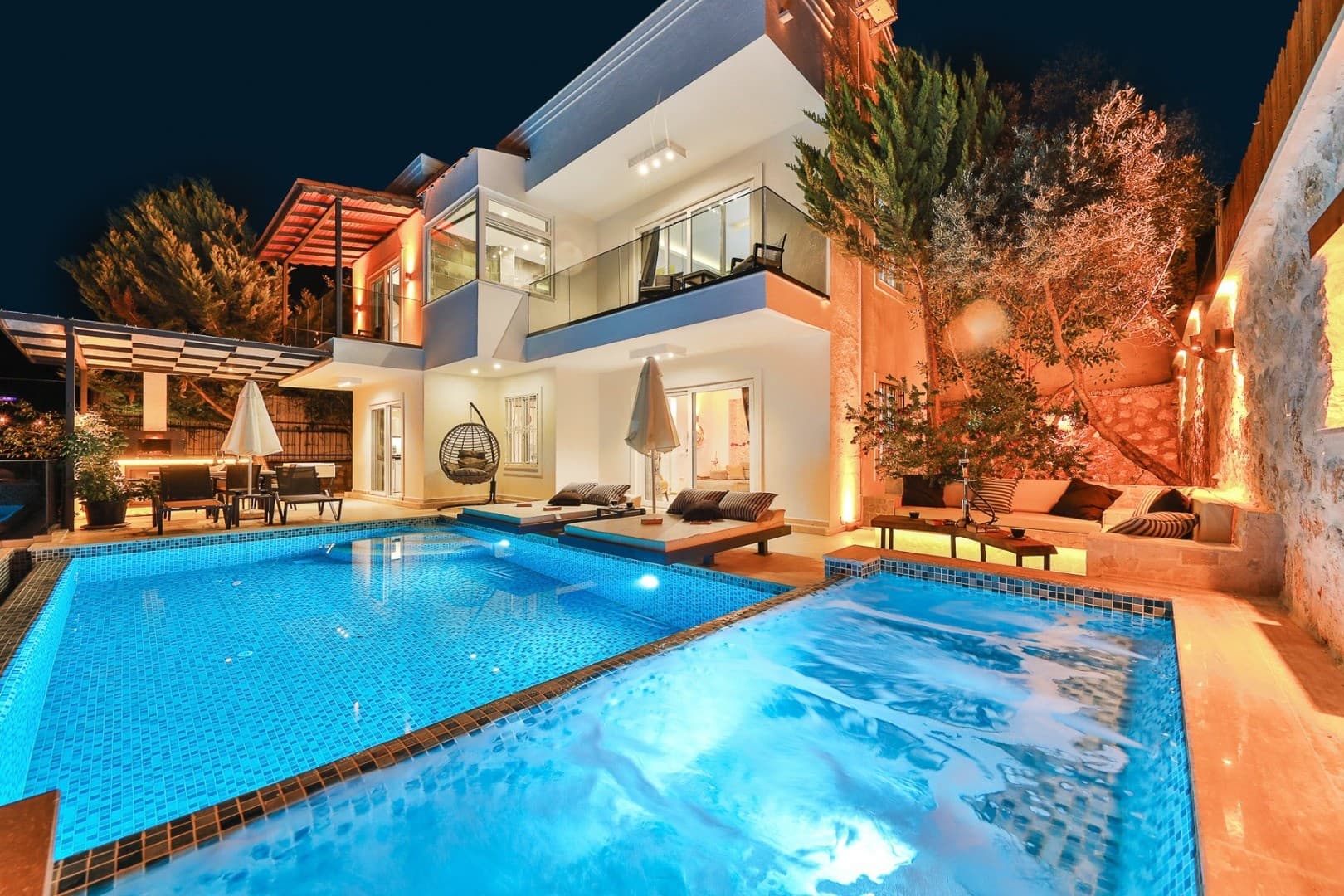 Gece aydınlatmalı, özel havuzlu ve şezlonglu iki katlı modern villanın bahçe görünümü.