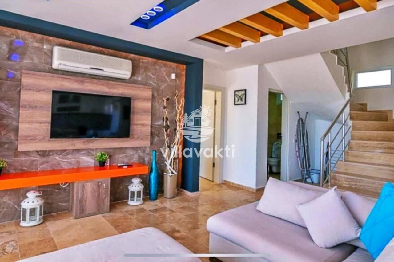 Bej koltuk takımı, duvara monte televizyon ve merdiven bulunan modern oturma odası.