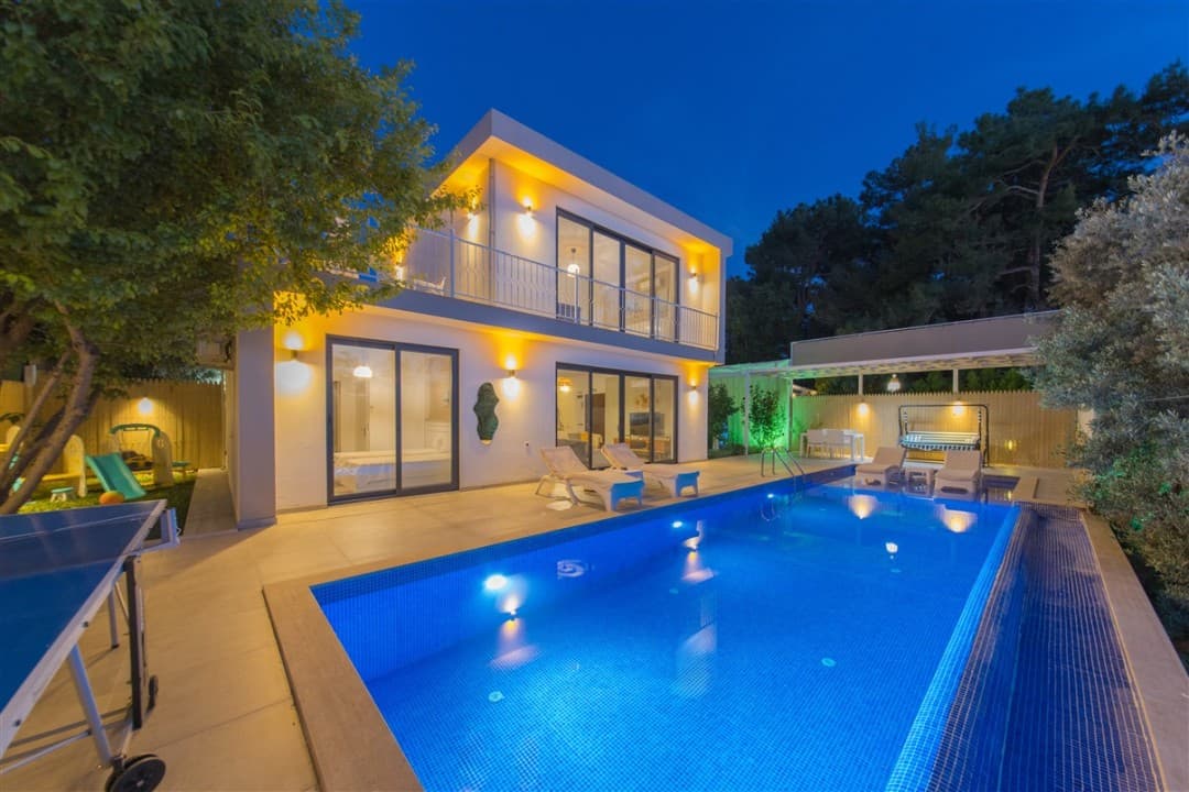 Gece aydınlatmalı havuzu, şezlongları ve masa tenisi bulunan iki katlı modern villa.