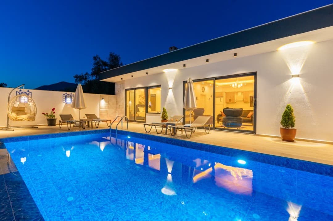 Gece aydınlatılmış havuzu ve şezlongları olan tek katlı beyaz villa.