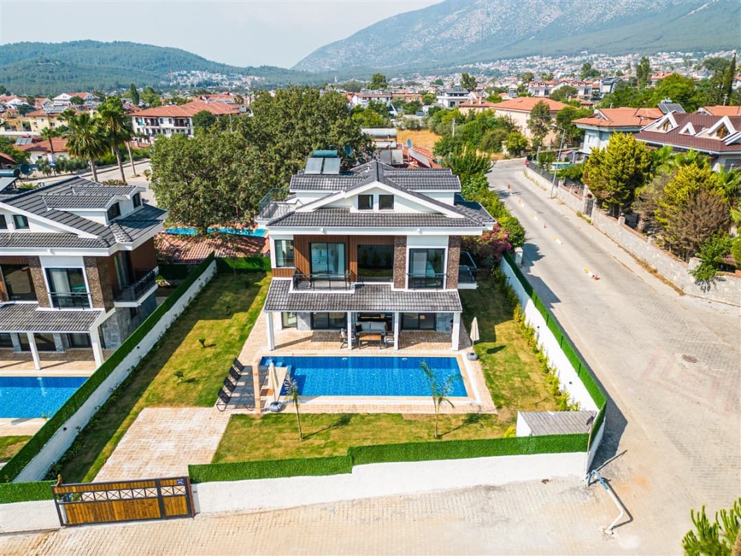 Üç katlı modern villa, önünde havuz, çim bahçe ve yanından geçen sokak.