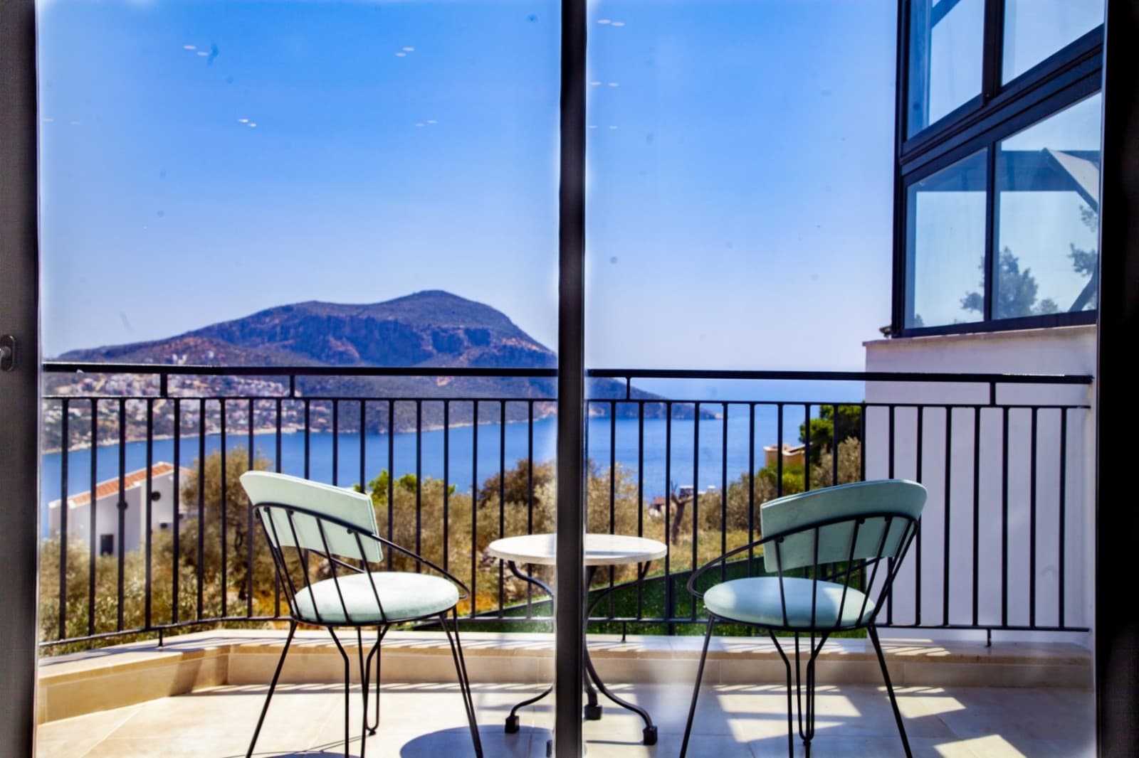 Balkon camının önünde iki açık renk sandalye ve sehpa, arka planda deniz ve dağ görünür.