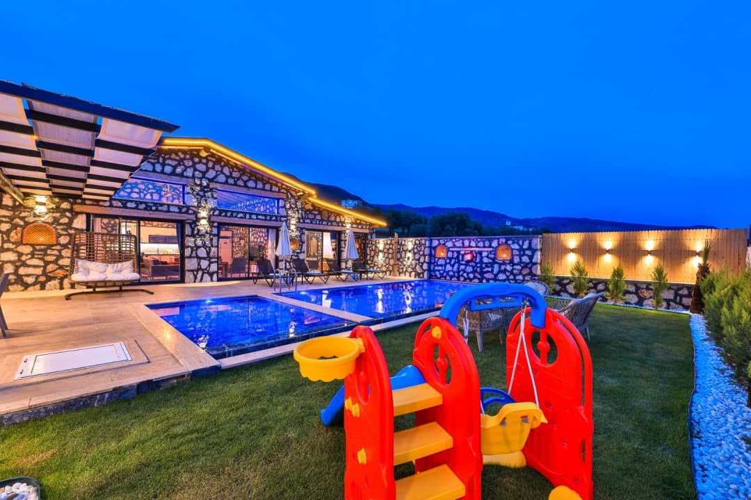 Akşam ışıklandırmalı taş villa, havuz ve çocuk oyun parkı.