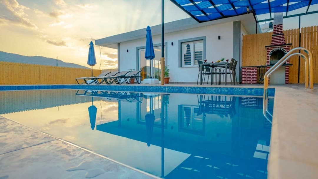Gün batımında havuz başı, şezlonglar, yemek masası ve tuğla barbekü alanı olan beyaz villa.