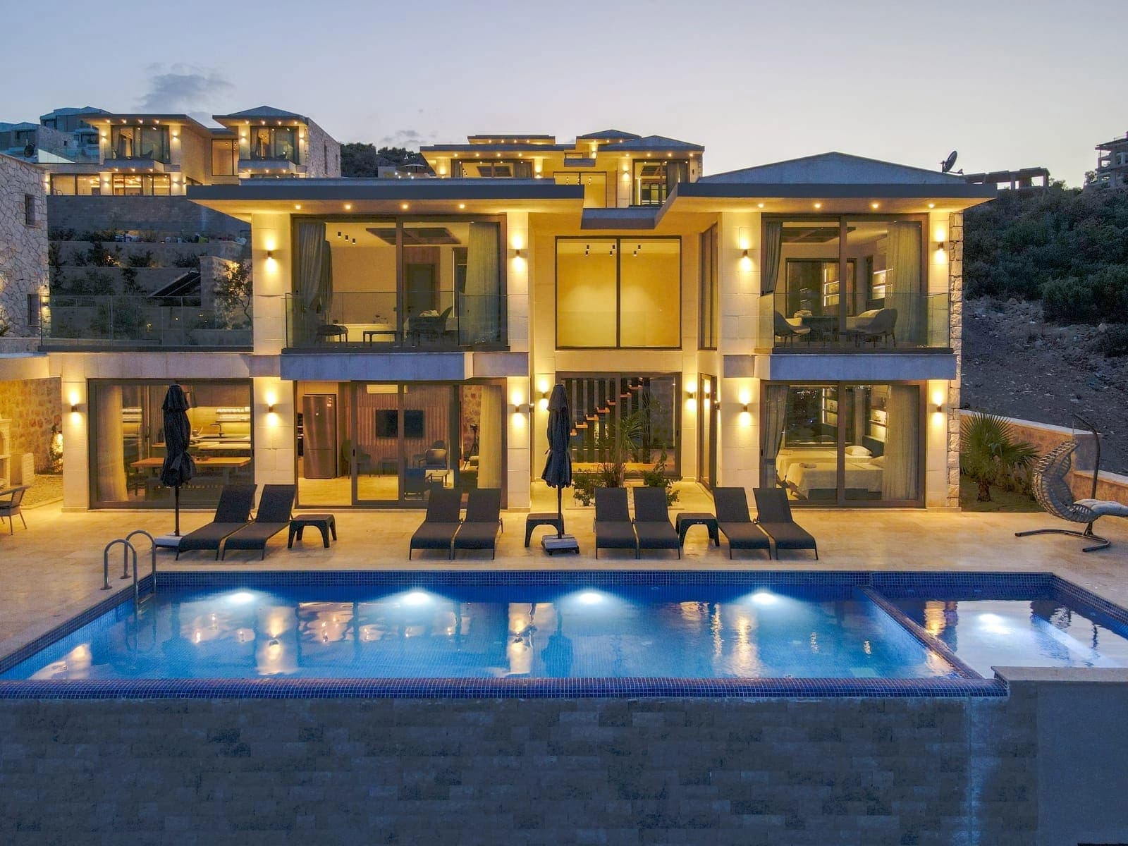 Akşam ışıklandırmalı havuz ve önünde şezlonglar bulunan iki katlı modern villanın dış cephesi.