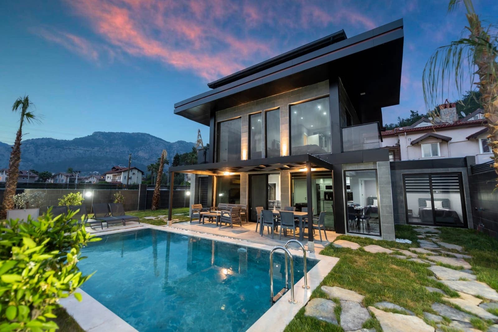 Dağ manzarası önünde konumlanan, havuzlu ve akşam ışıklandırılmış modern villanın dış cephesi.