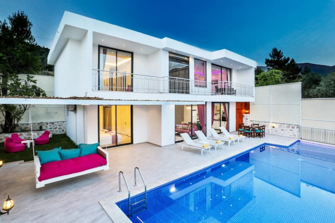 Akşam vakti aydınlatılmış, özel havuzlu, beyaz dış cepheli iki katlı villa ve bahçesinde oturma grubu.