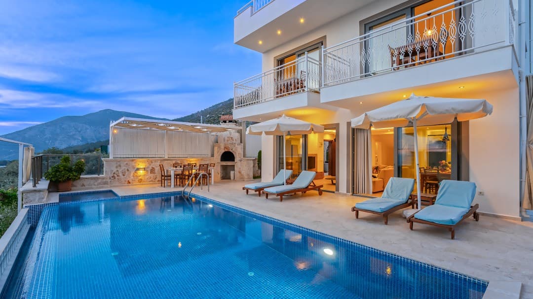 Akşam vakti aydınlatılmış, havuz başında şezlonglar, güneş şemsiyeleri ve taş barbekü bulunan iki katlı villa.