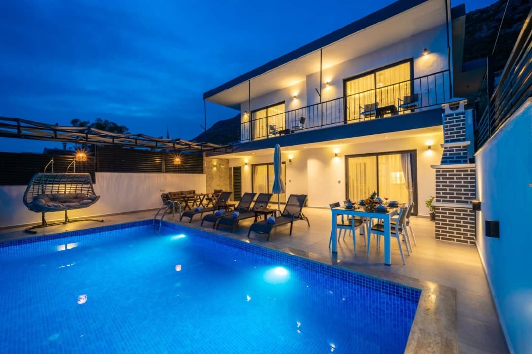 Akşam vakti havuz başı şezlongları ve yemek masası olan iki katlı aydınlatılmış villa.