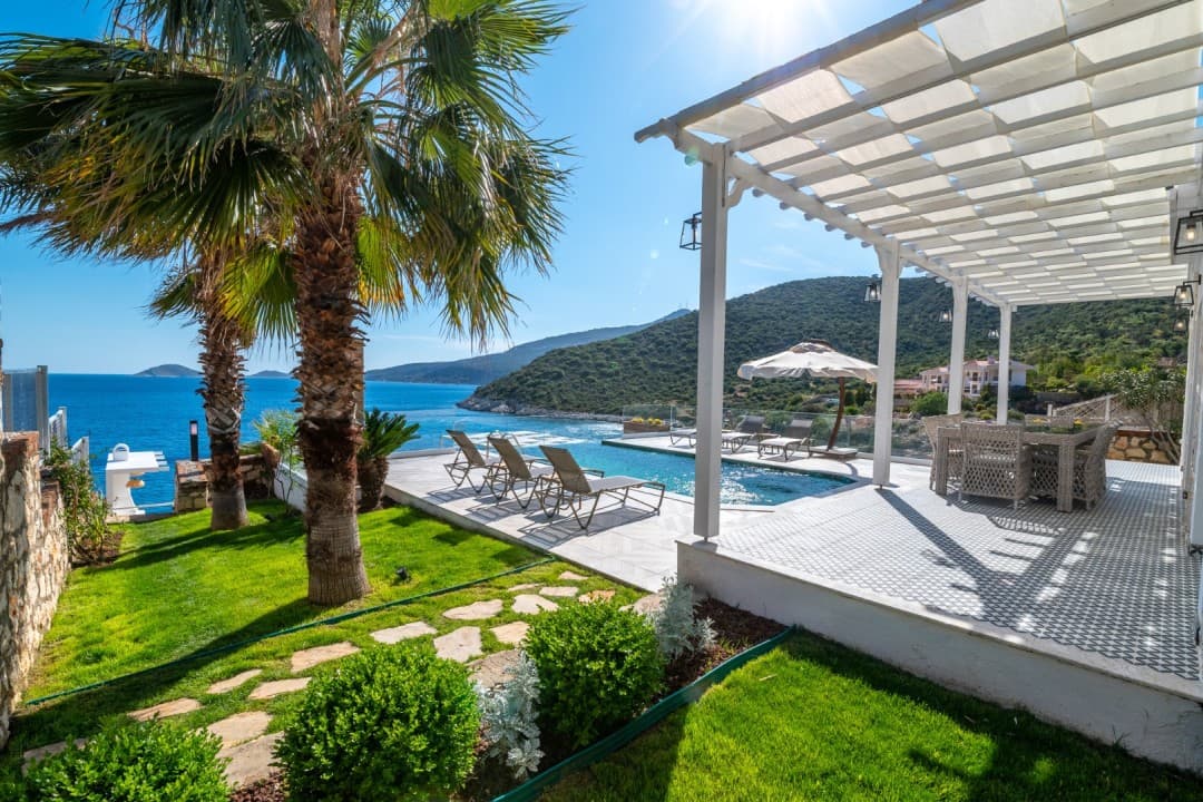 Deniz manzaralı havuz başı terasında şezlonglar, palmiyeler ve beyaz pergola yer alıyor.
