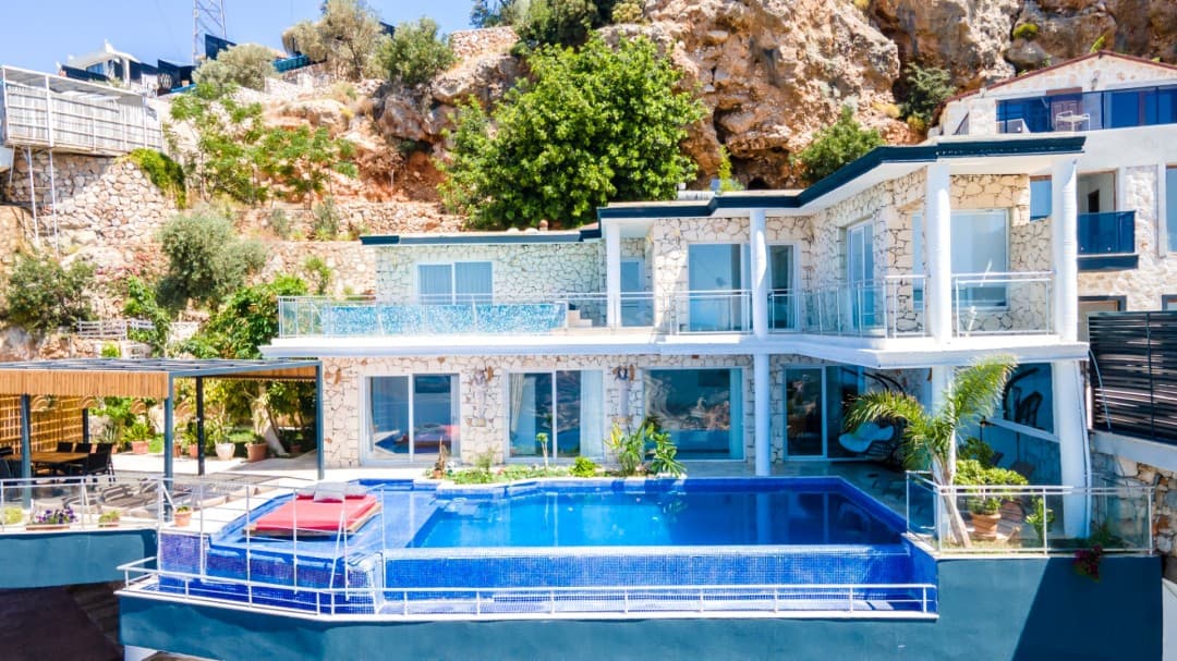 Taş kaplama cepheli, önünde mavi havuz ve şezlong bulunan villa.