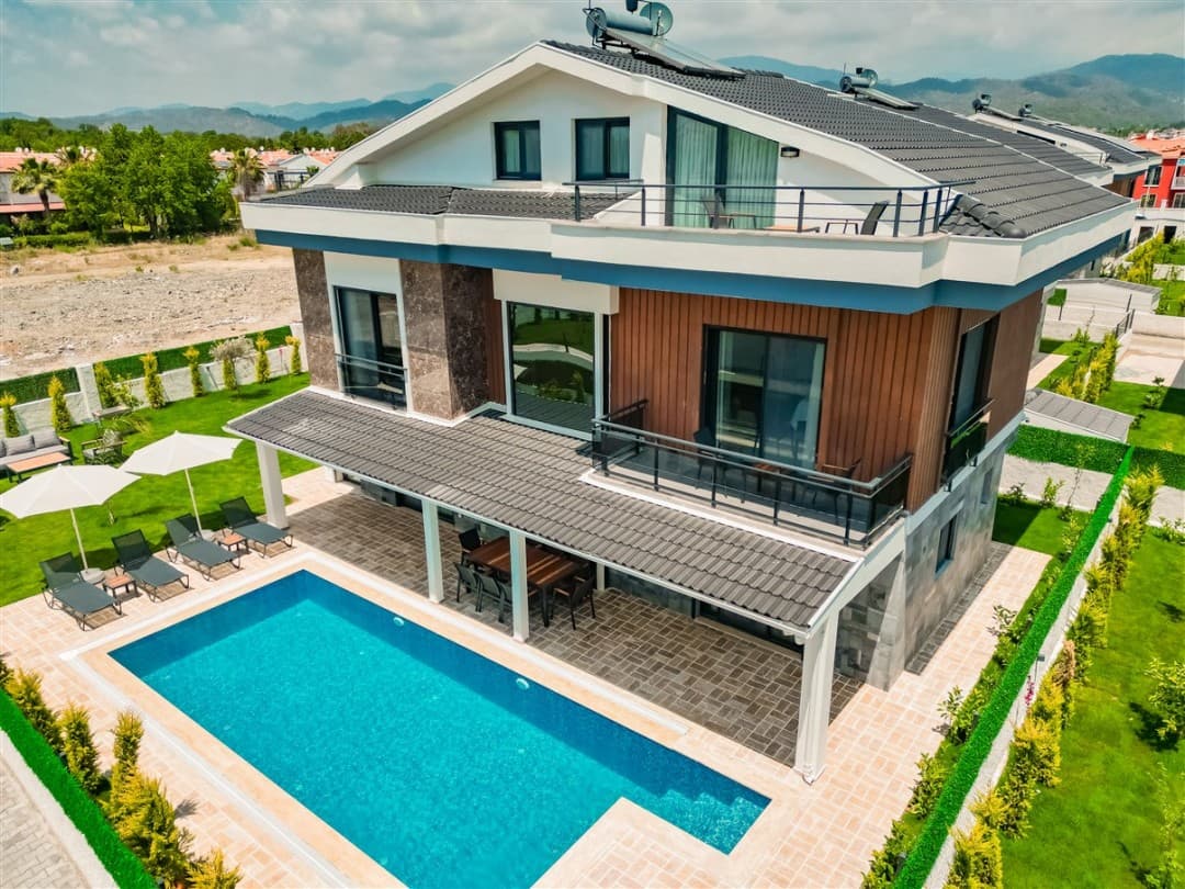 Havuz kenarında şezlonglar ve beyaz şemsiyeler bulunan, dağ manzaralı modern villa dış görünümü.