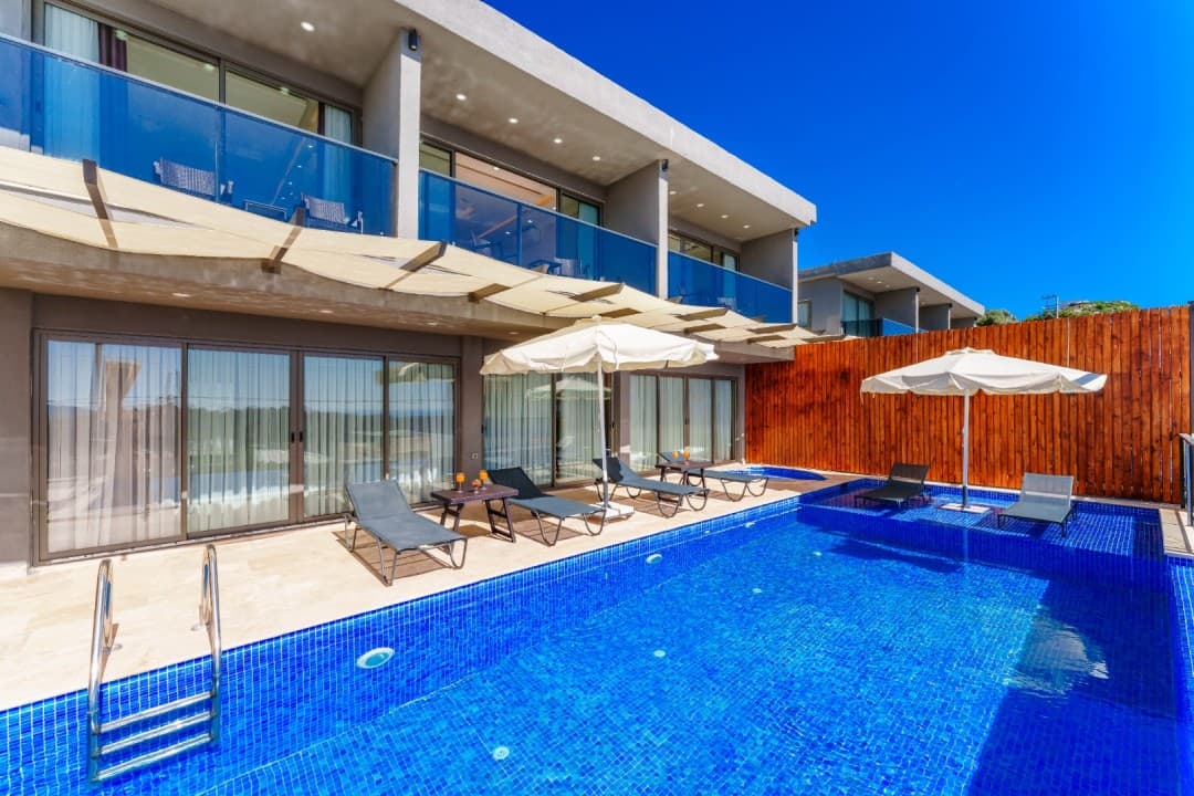 Gündüz saatlerinde havuz başında şezlonglar ve şemsiyeler bulunan iki katlı villa.