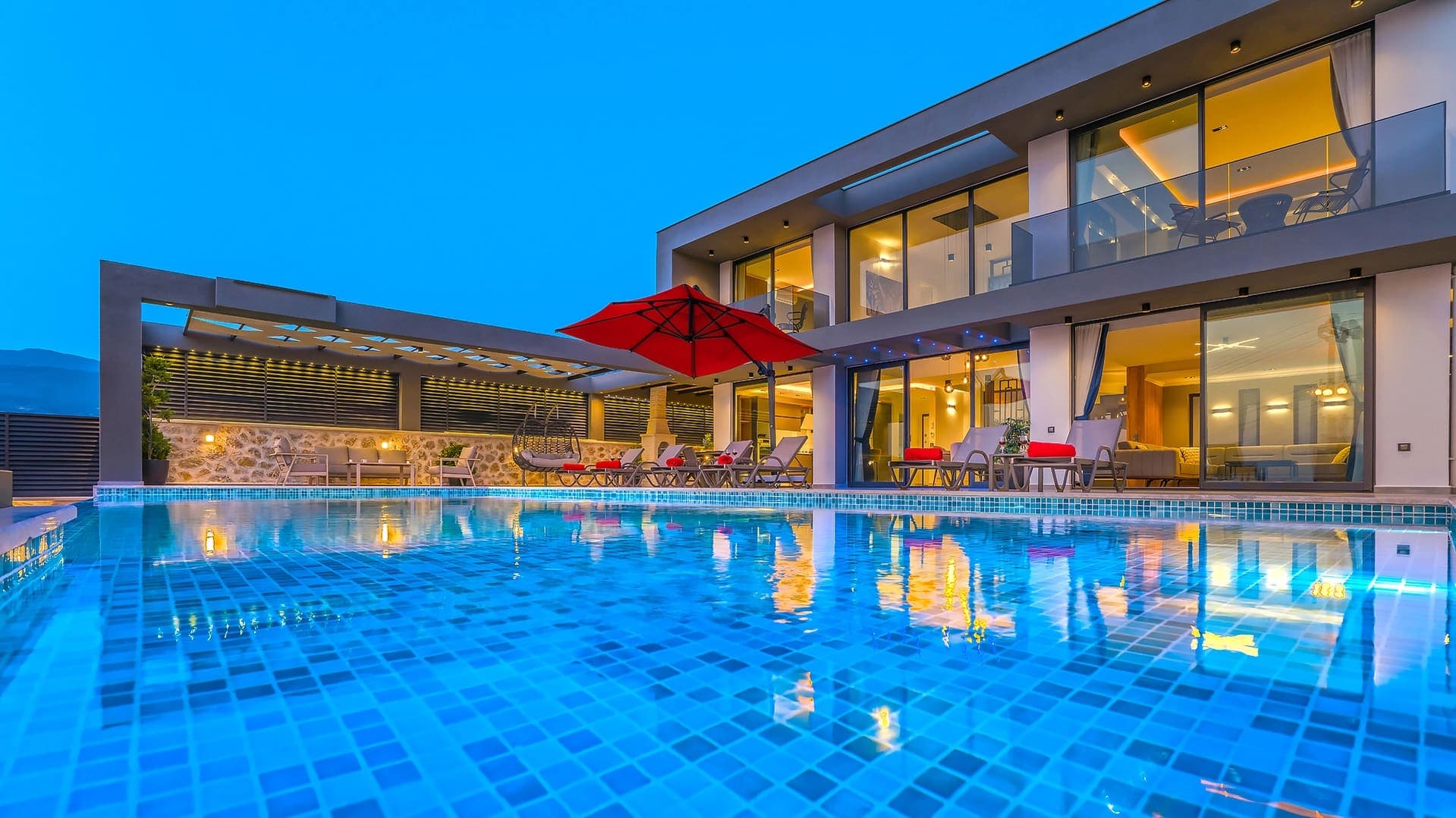 Akşam karanlığında havuz başı şezlongları ve aydınlatmalı modern villanın dış görünümü.