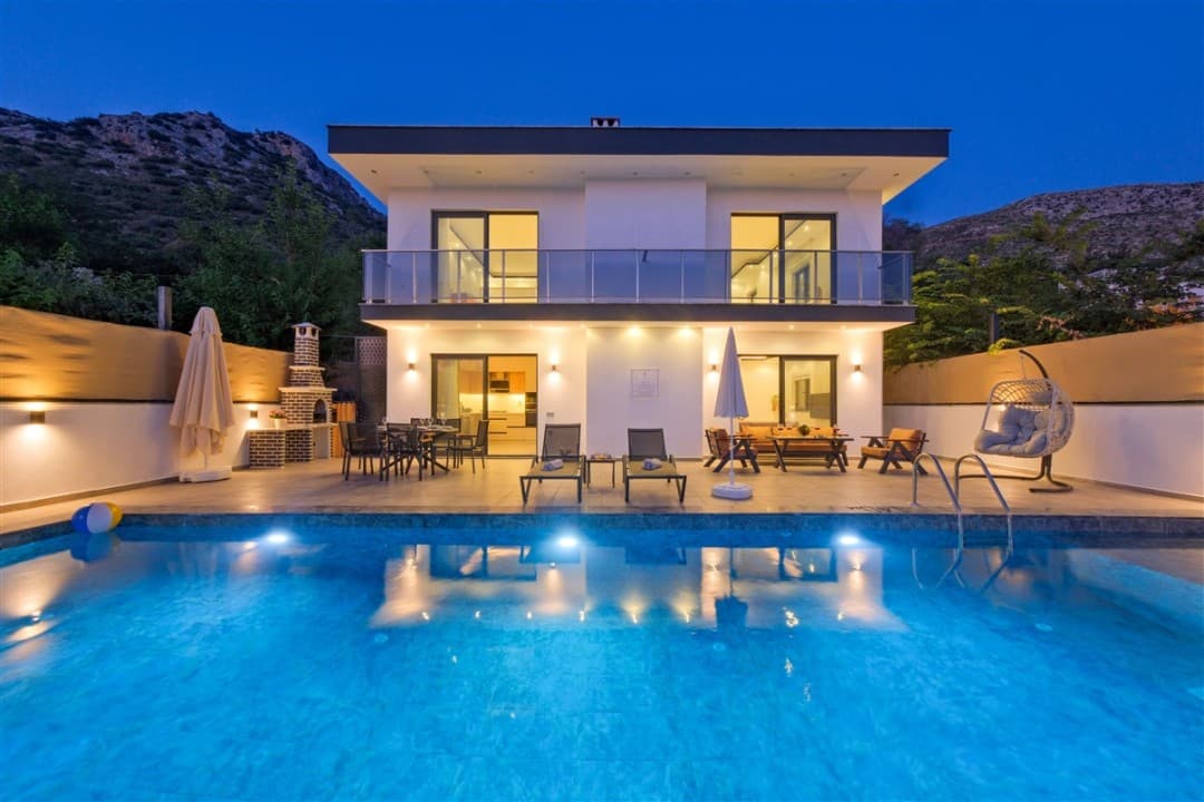 Aydınlatmalı havuz kenarında şezlonglar bulunan iki katlı villa.