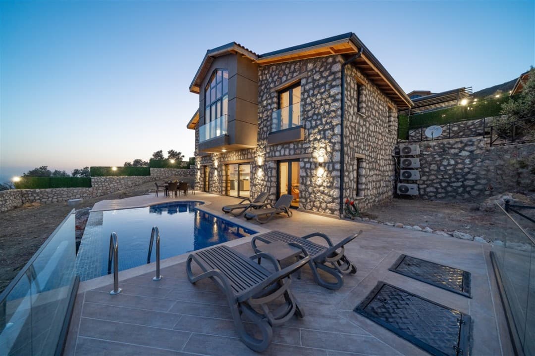 Alacakaranlıkta taş duvarlı, havuzlu ve şezlonglu iki katlı villa.