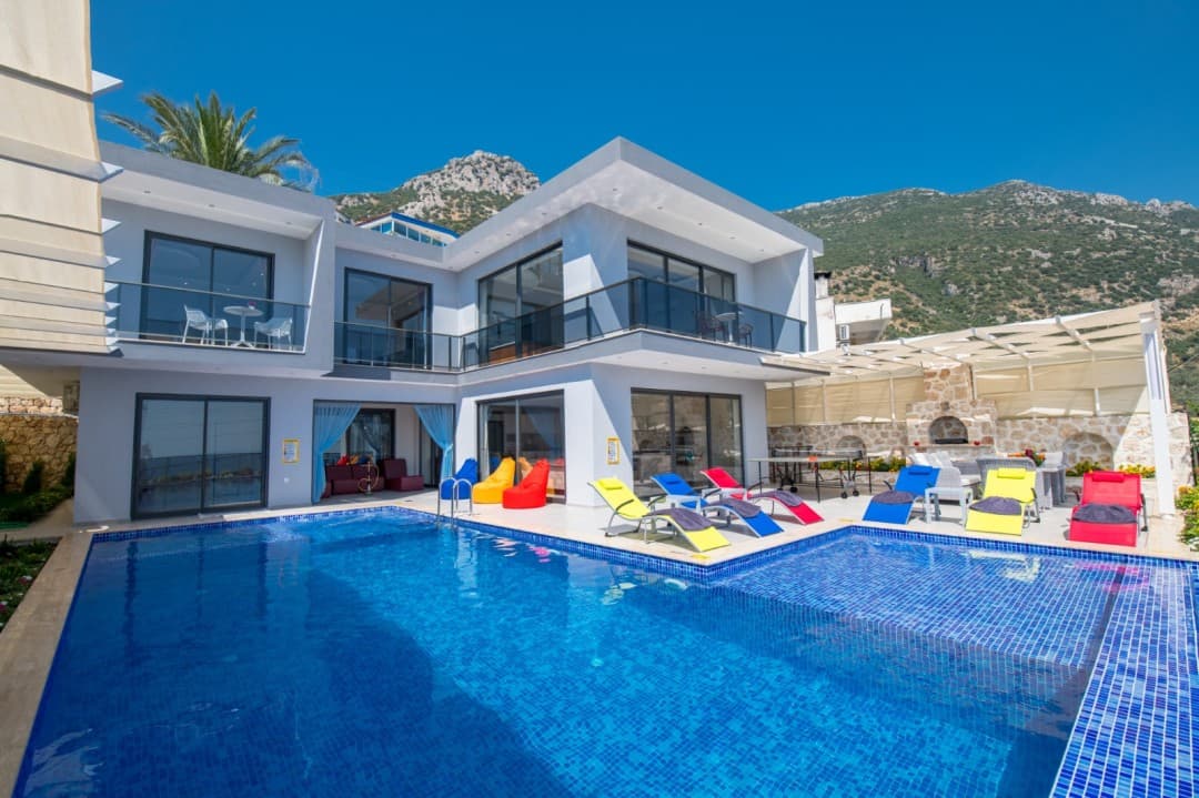Dağ manzaralı, iki katlı modern villanın önünde mavi bir yüzme havuzu ve renkli şezlonglar bulunuyor.