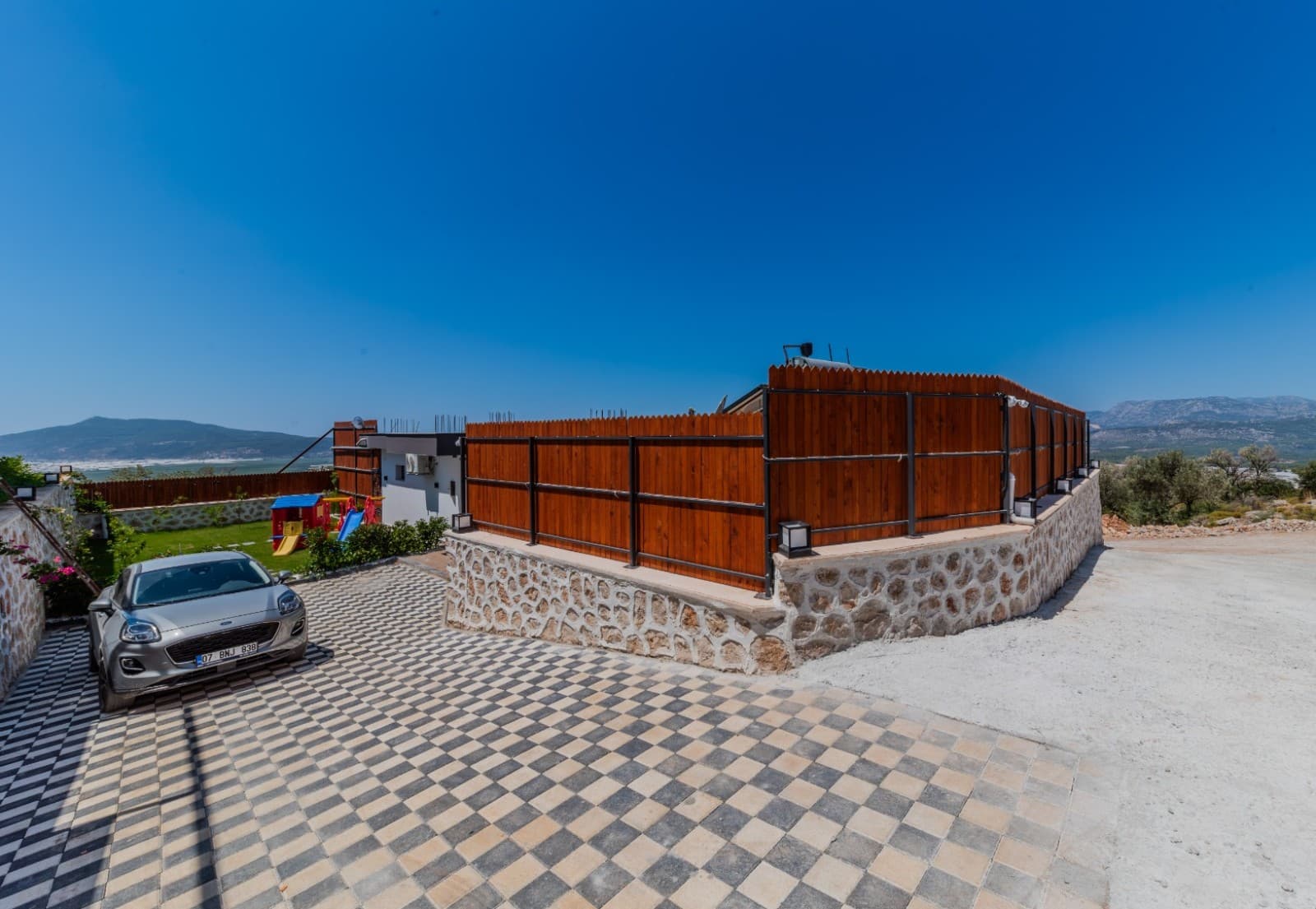 Taş duvarlı ve ahşap çitli otopark alanında park edilmiş gri otomobil.