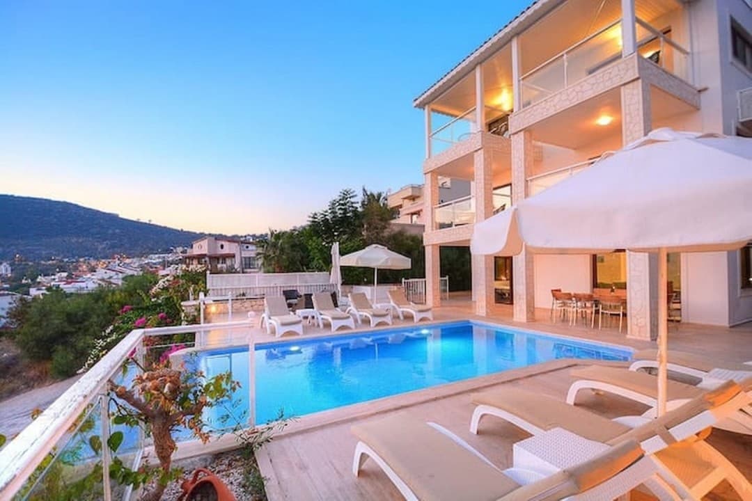 Akşam vakti havuz başında şezlonglar, güneş şemsiyeleri ve balkonlu üç katlı bir villa.