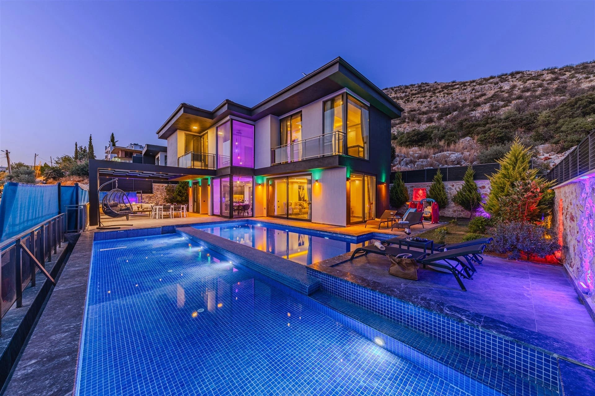 Akşam vakti renkli ışıklarla aydınlatılan havuzlu modern villanın dış cephesi.