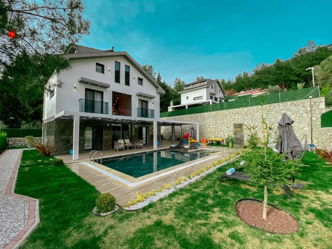 Yeşil bahçesi, havuzu ve ağaçlarla çevrili taş duvarları olan beyaz dış cepheli villa.
