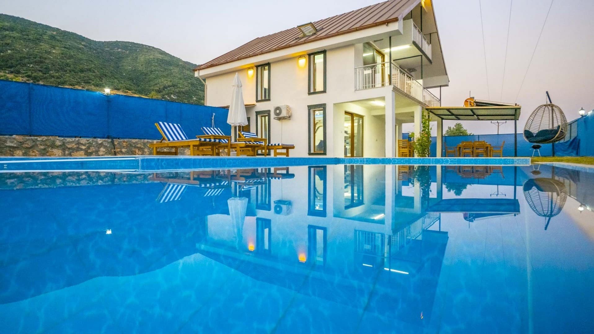 Ön planda yüzme havuzu, arkasında beyaz dış cepheli iki katlı villa, şezlonglar ve dağ manzarası.