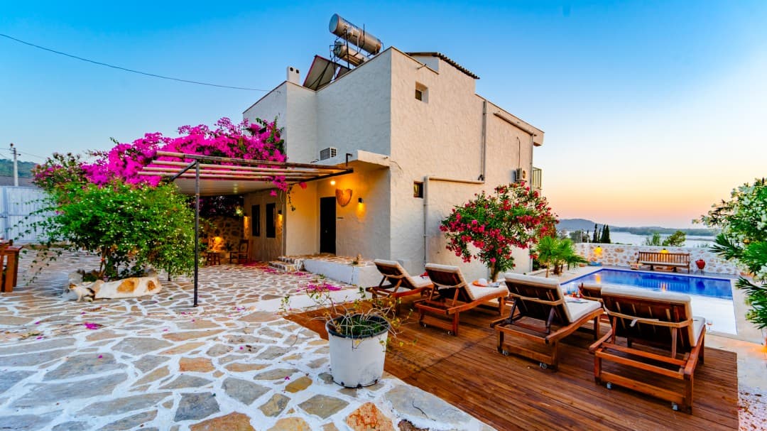 Pembe begonvillerle çevrili, havuzlu ve ahşap şezlonglu bahçeye sahip beyaz dış cepheli müstakil villa.
