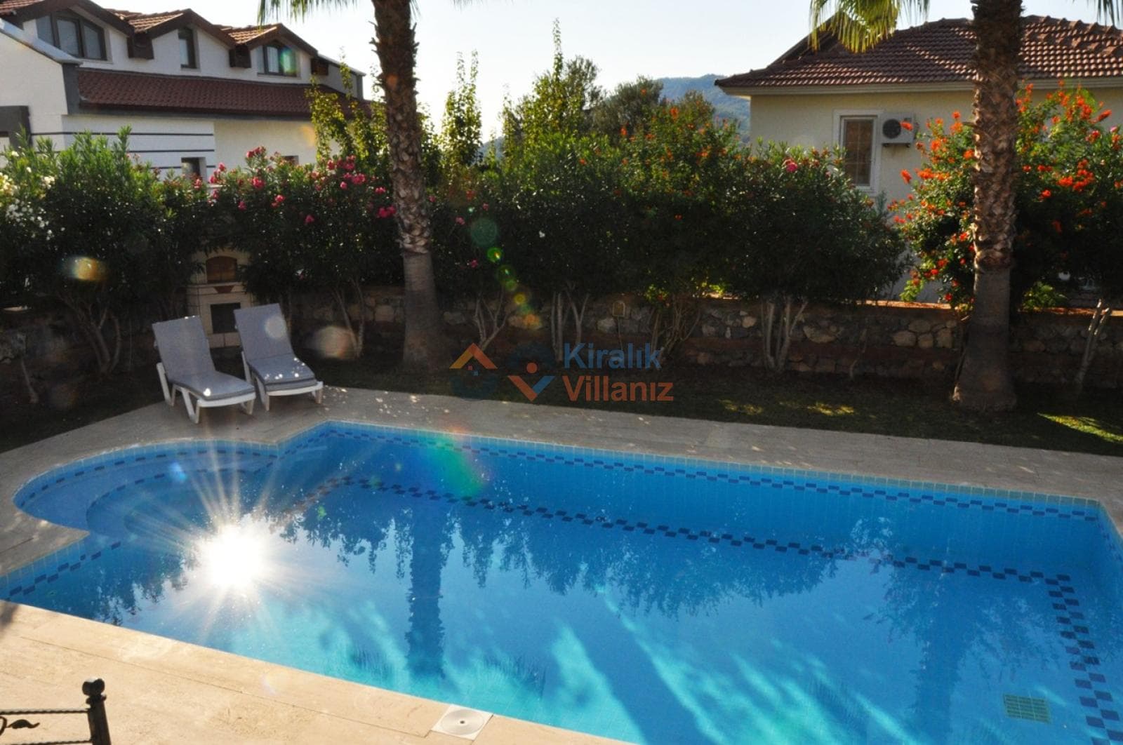 Bahçesinde havuz ve şezlonglar bulunan, çiçeklerle çevrili villa dış mekanı.