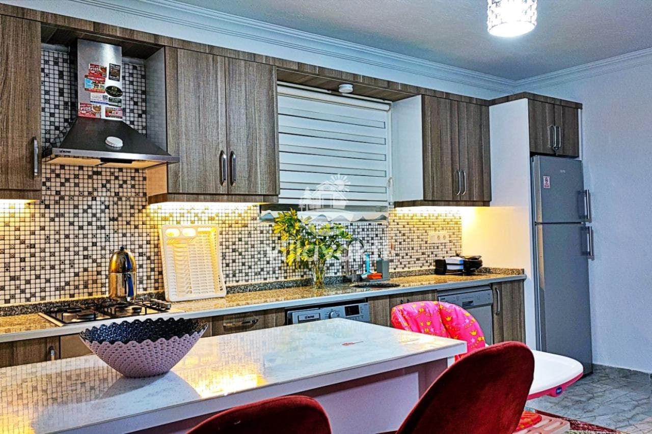 Ahşap dolaplı ve tezgahlı modern mutfakta gri buzdolabı ve yemek masası var.