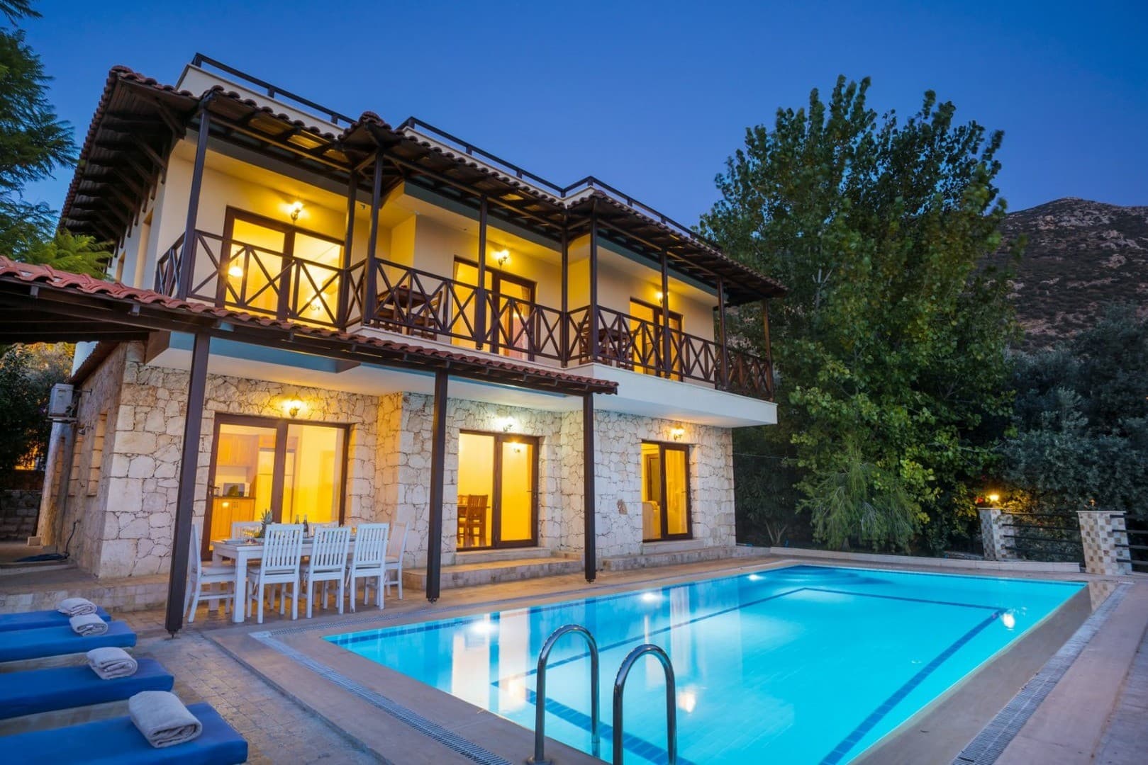 Akşam vakti aydınlatılmış, taş kaplama dış cepheli, havuz başında yemek masası bulunan iki katlı villa.
