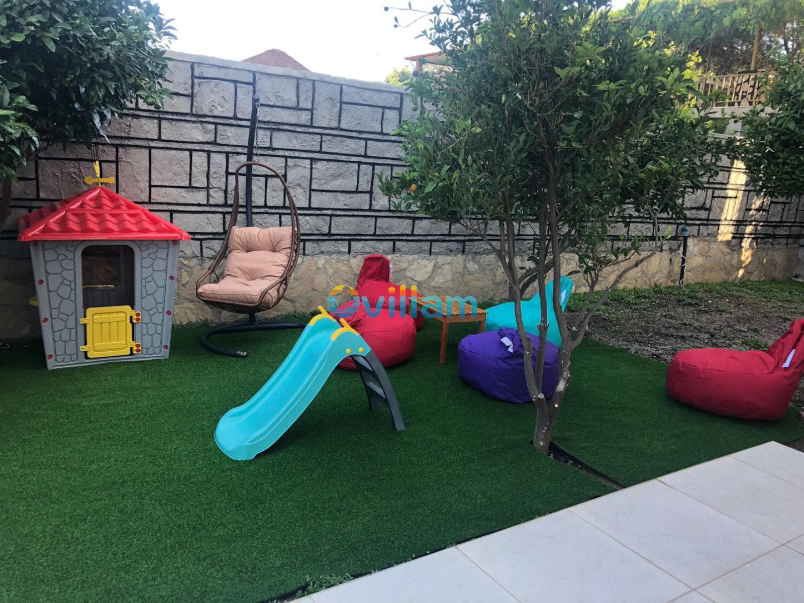 Çim zeminli bahçede çocuk oyun evi, kaydırak ve minderli oturma alanları