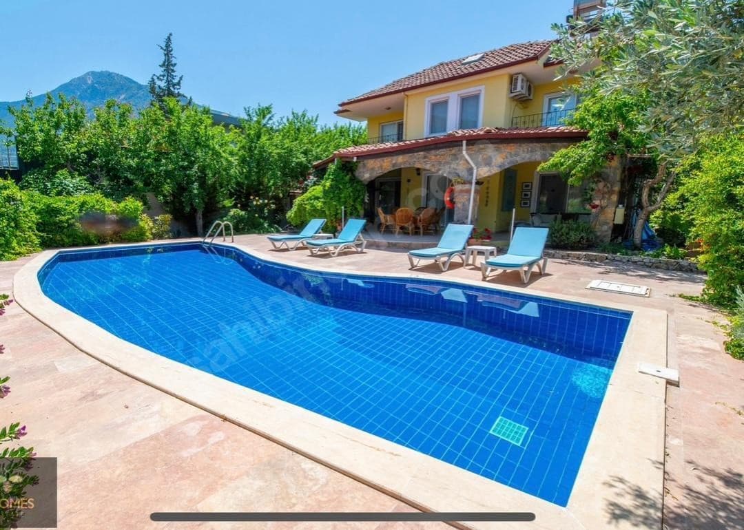 Önünde mavi yüzme havuzu ve şezlonglar bulunan, taş detaylı, sarı renkli iki katlı müstakil villa.