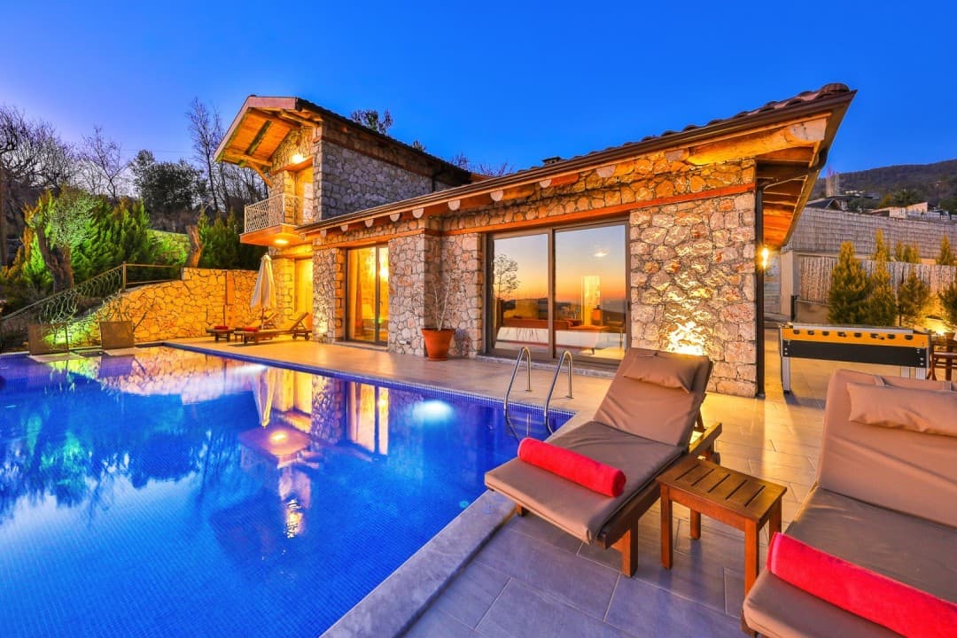 Alacakaranlıkta taş cepheli villa, havuz başında şezlonglar ve masa tenisi.