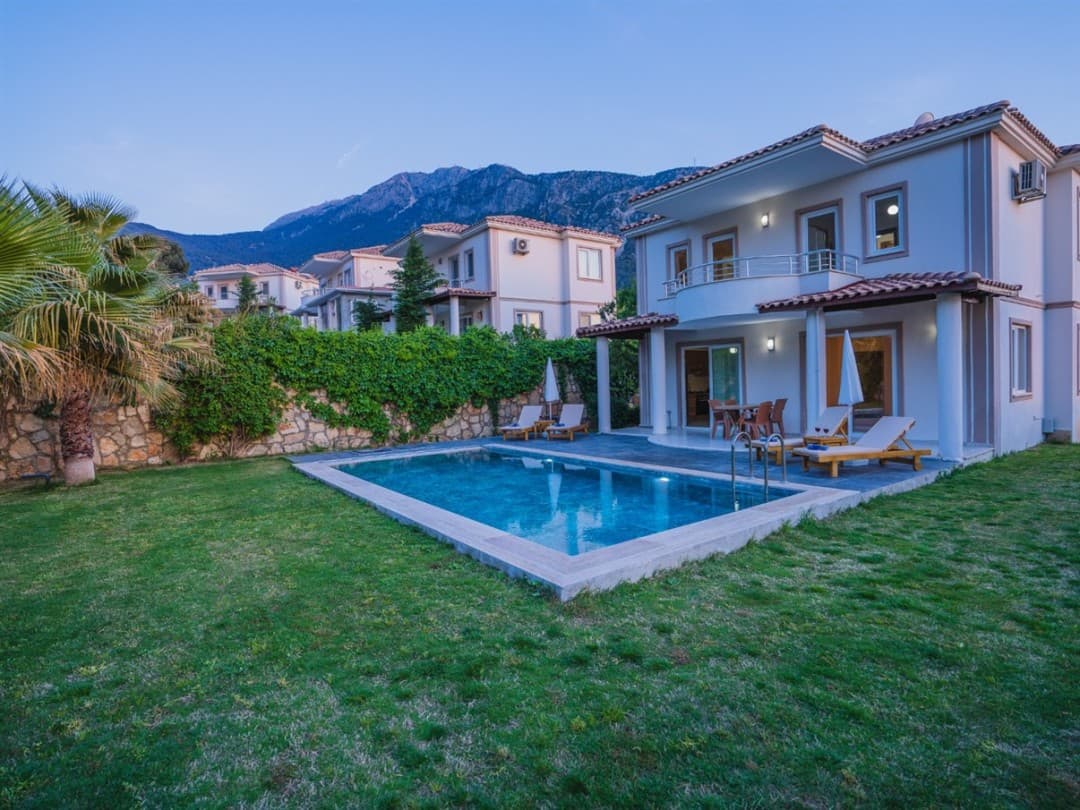 Bahçesinde havuz ve şezlonglar bulunan iki katlı villa ve dağ manzarası.