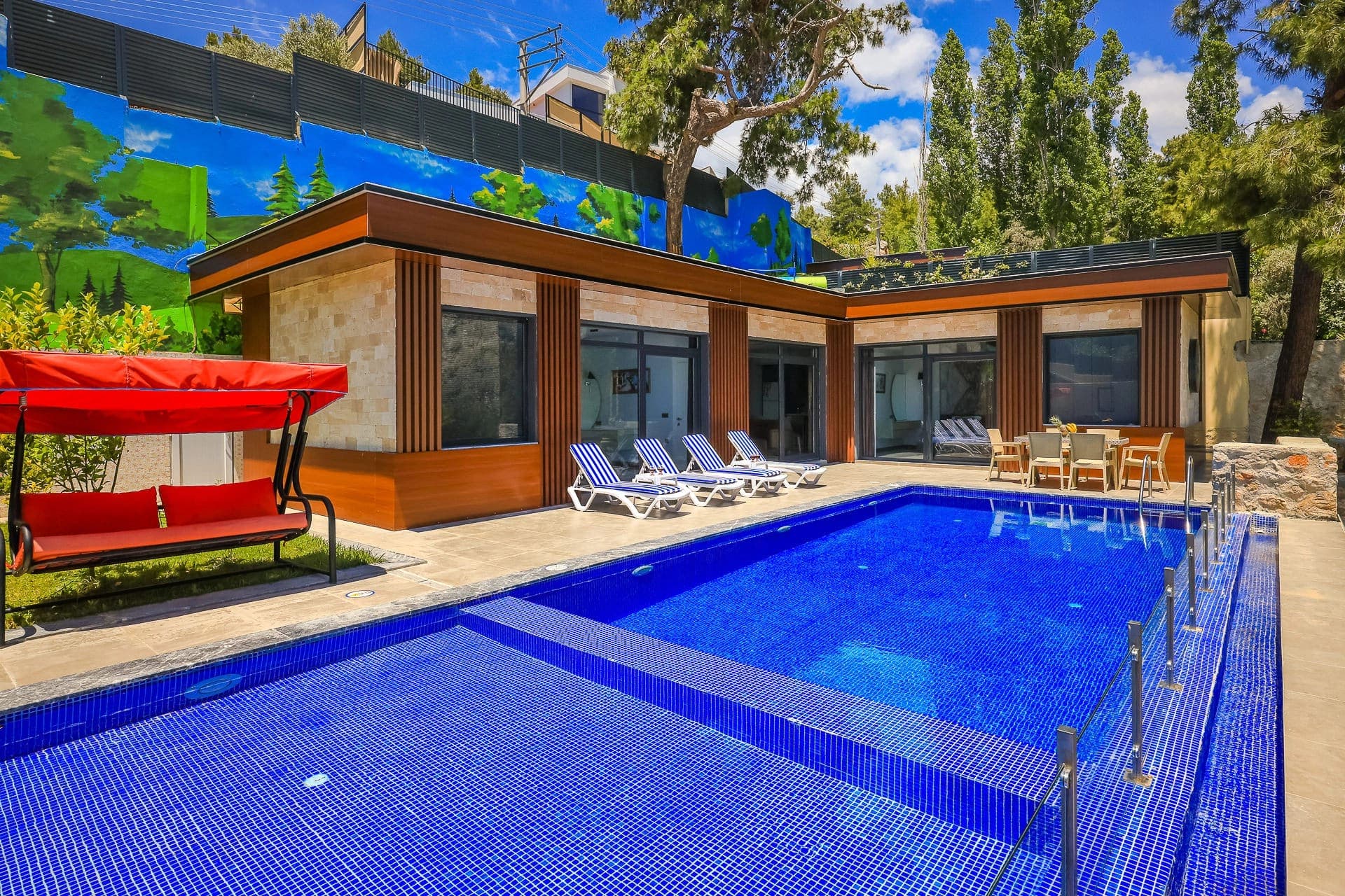 Önünde mavi havuz ve kırmızı salıncak bulunan tek katlı modern villanın dış cephesi.