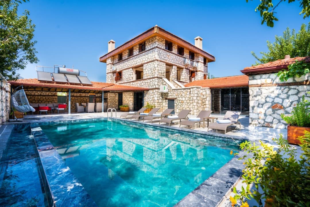 Önünde havuz ve şezlonglar bulunan, taş duvarlı üç katlı villa.