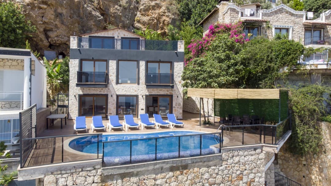 Kayalık yamaca kurulu, önünde havuz, şezlonglar ve kamelya bulunan taş ve koyu gri tonlu iki katlı villa.
