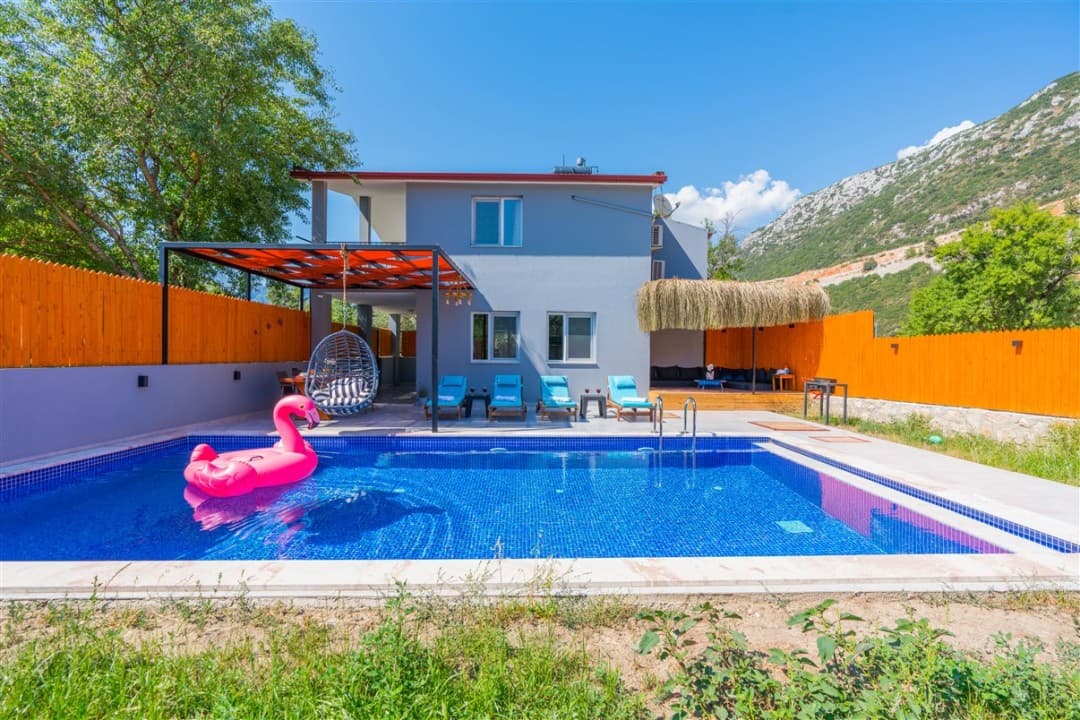 Önünde pembe flamingo deniz yatağı olan havuzlu, ahşap çitlerle çevrili bahçeli villa ve dağ manzarası.