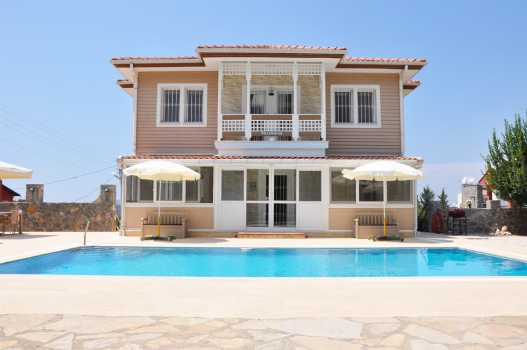 Önünde yüzme havuzu, beyaz şemsiyeler ve ahşap banklar bulunan iki katlı, bej renkli müstakil villa.