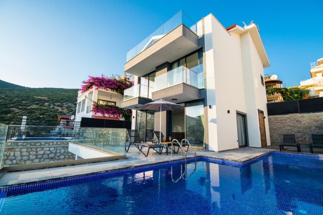Önünde özel yüzme havuzu, şezlonglar ve güneş şemsiyesi bulunan modern mimarili beyaz bir villa.