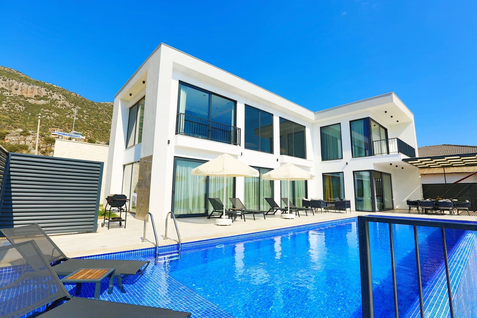 Mavi mozaikli yüzme havuzunun arkasında iki katlı modern beyaz villa.