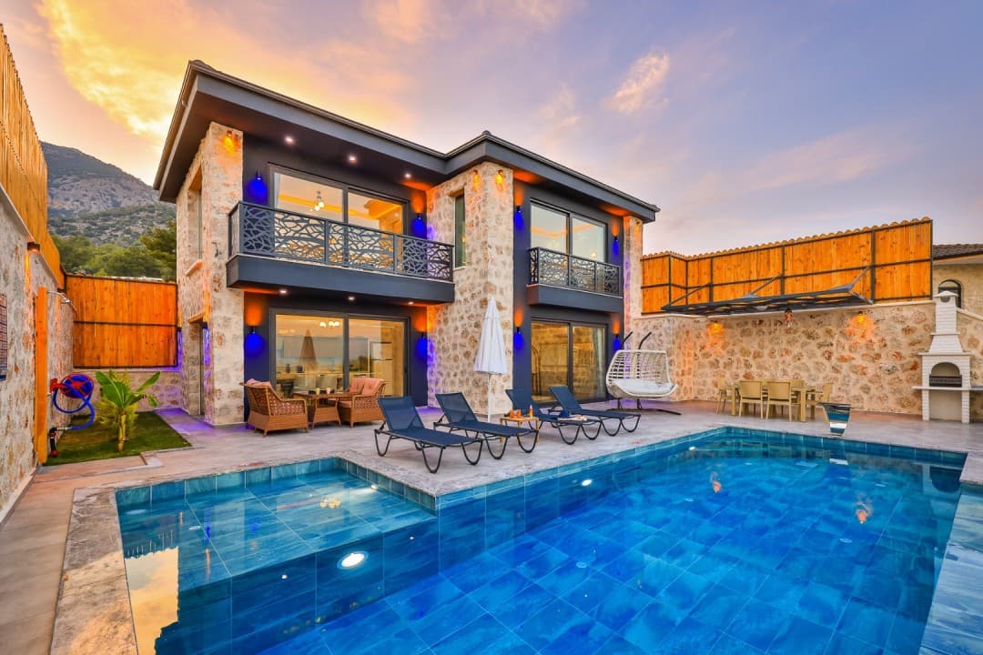 Gün batımında önünde havuz, şezlonglar ve taş duvarlı iki katlı villa.