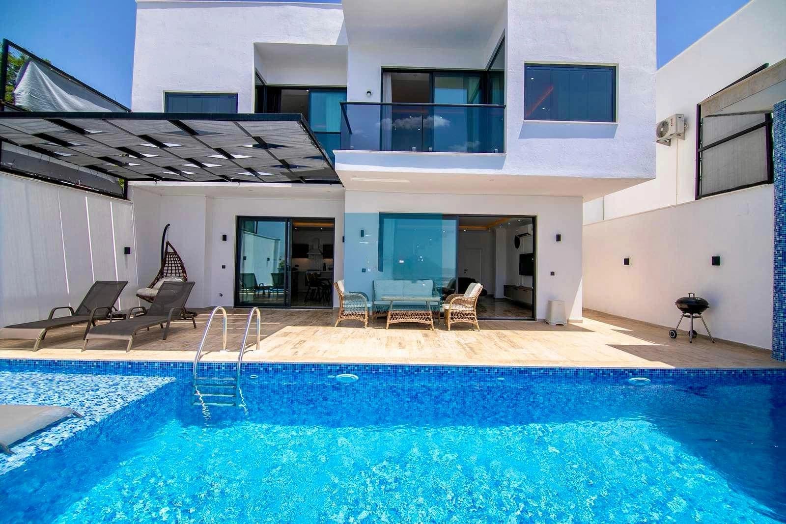 Modern beyaz villa, önünde mavi yüzme havuzu, şezlonglar, oturma grubu ve barbekü alanı yer alıyor.