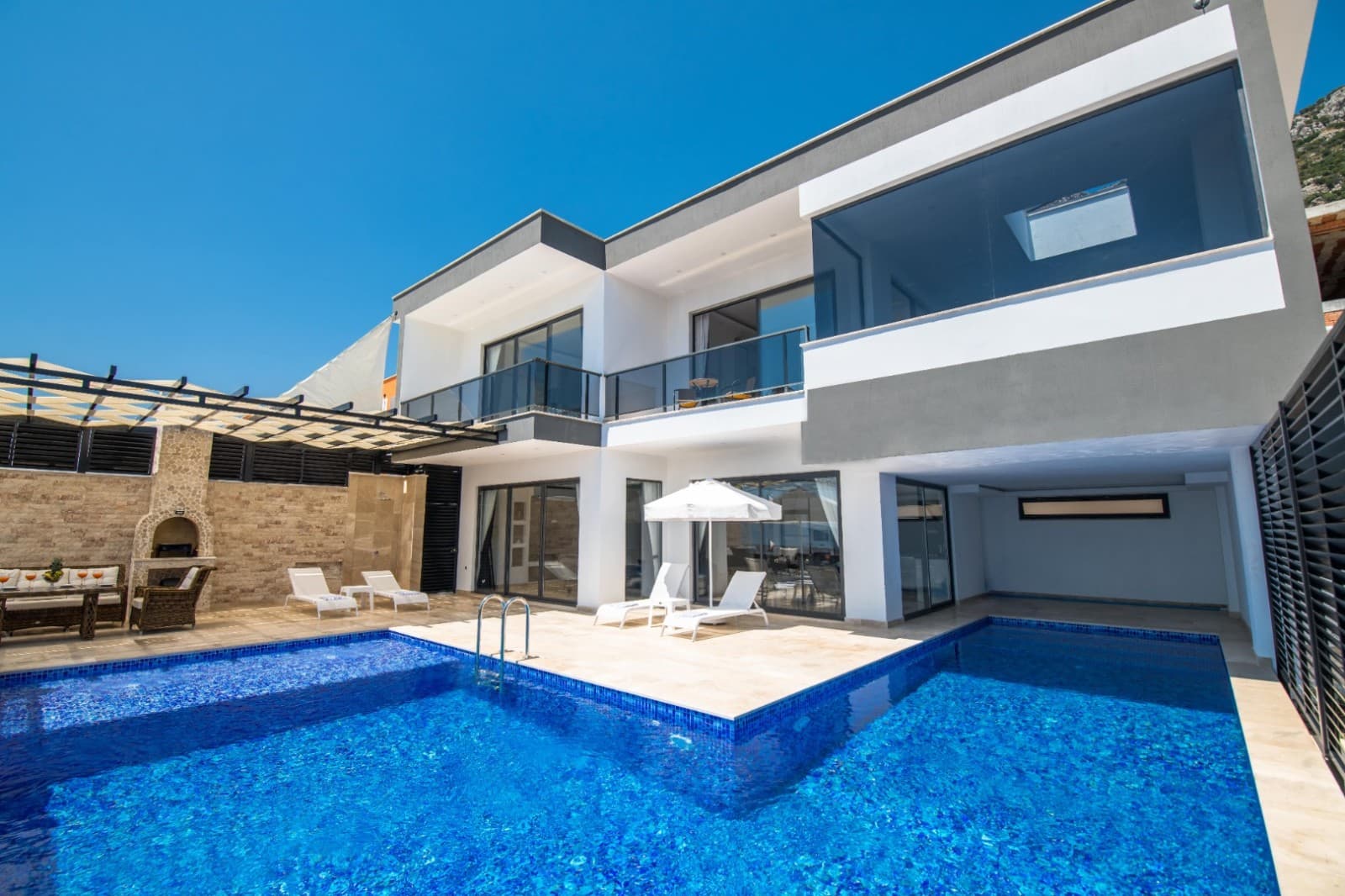 Gündüz açık mavi gökyüzü altında iki katlı modern villa ve önündeki mavi havuz.