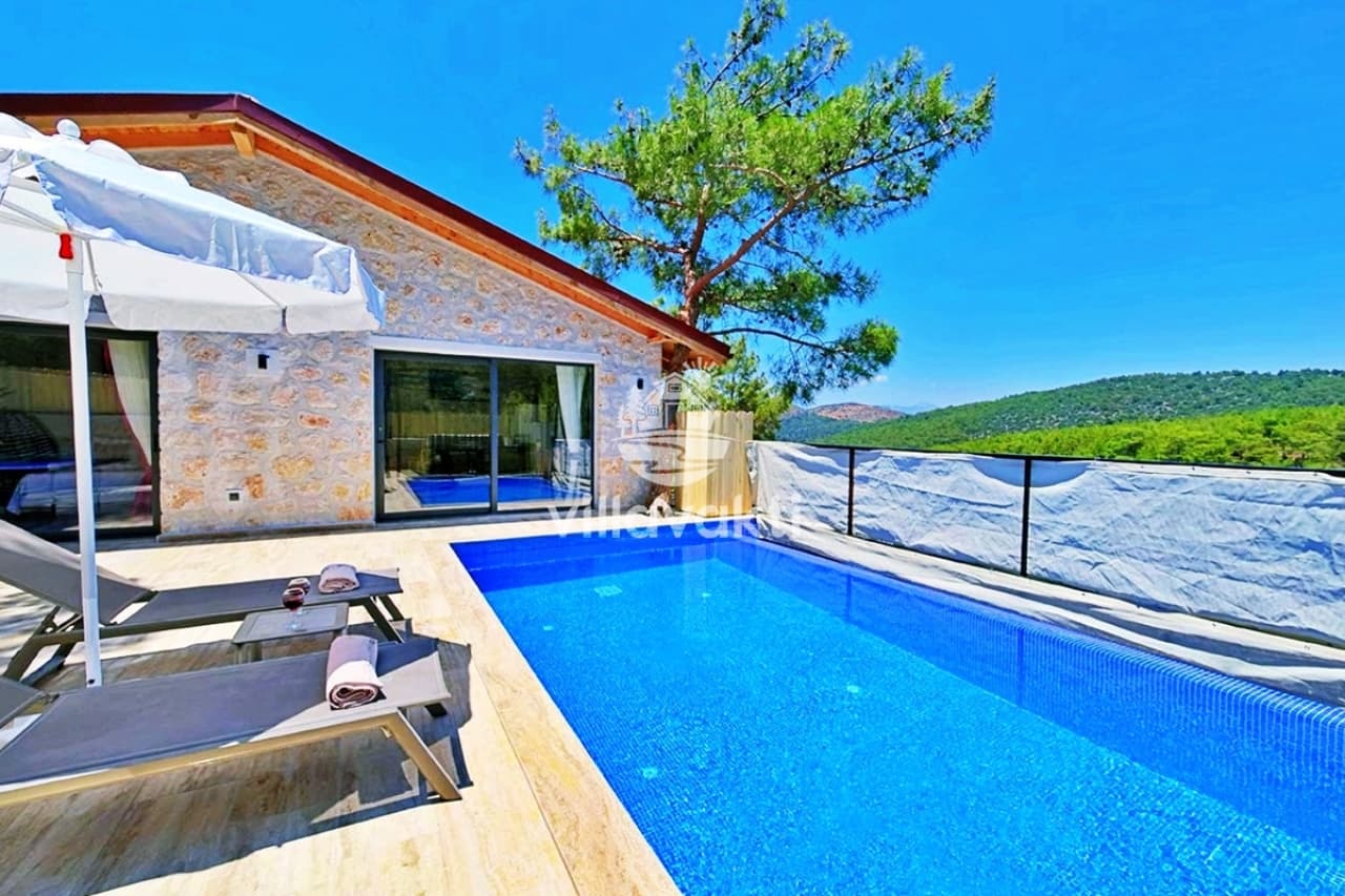 Taş duvarlı villa önünde güneş şemsiyesi, şezlonglar ve mavi bir havuz ile orman manzarası.