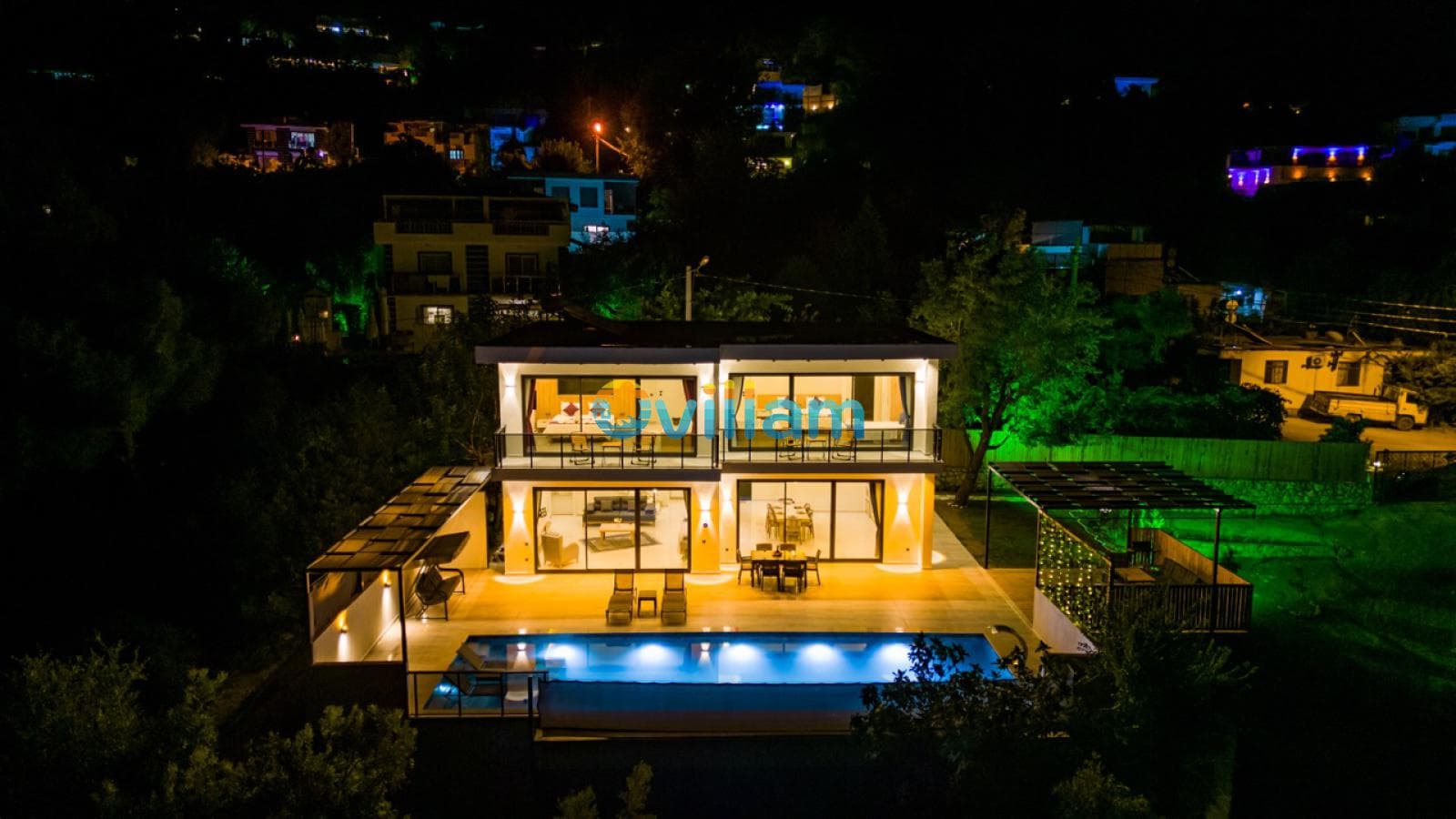 Gece ışıklandırılmış iki katlı villa, önündeki yüzme havuzu ve çevresindeki doğa manzarası.