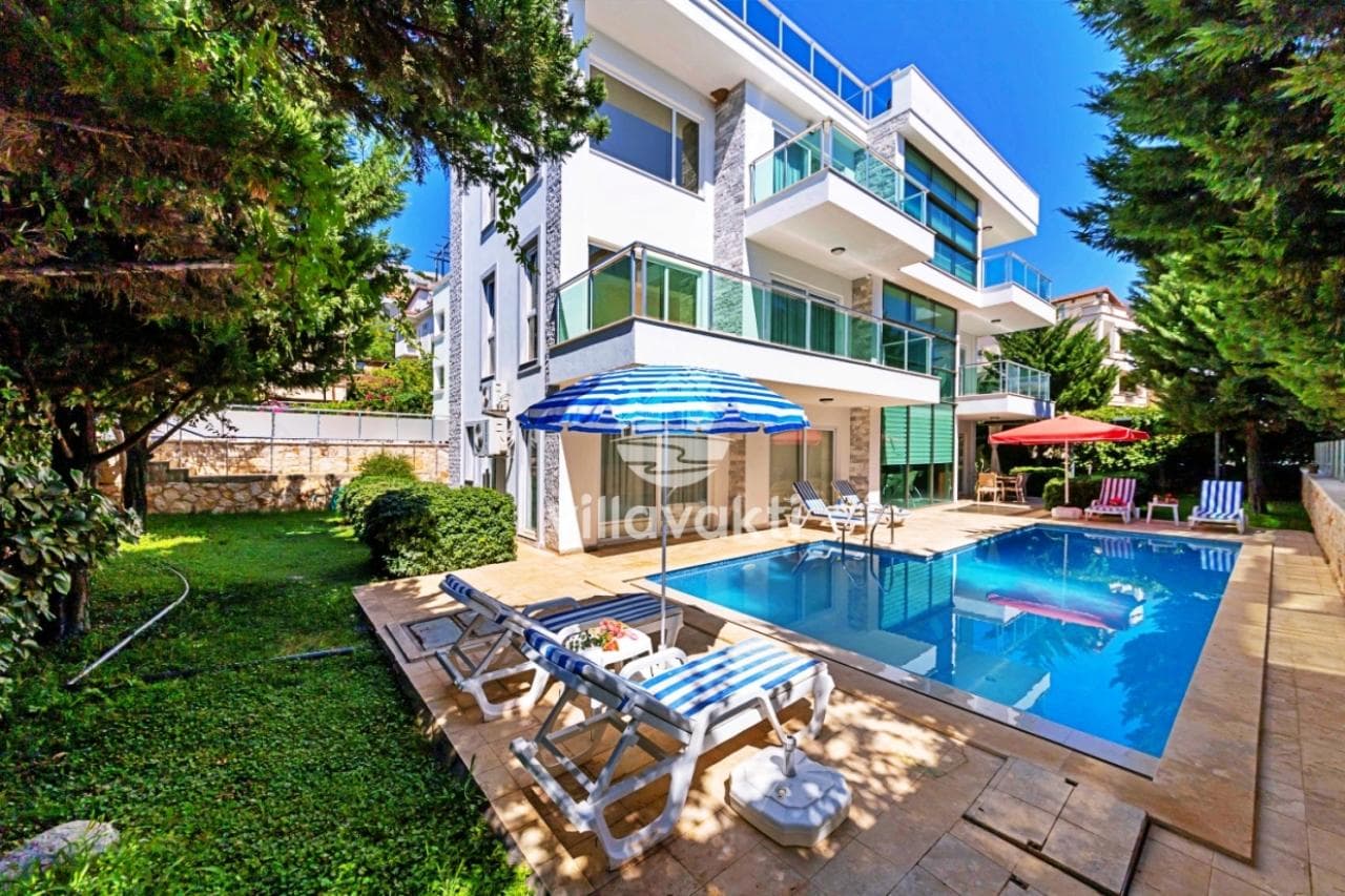 Mavi havuzlu, şezlonglu ve yeşil bahçeli villa dış cephesi.