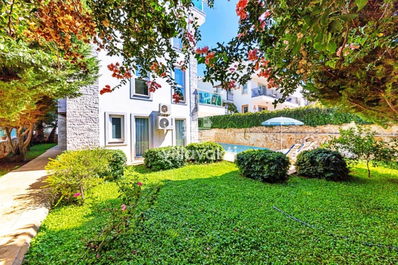 Yeşil çimler ve çiçekli ağaçlar arasında villa dış cephesi.